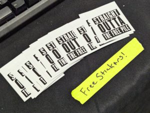 Stack of black-and-white stickers reading 'Straight Outta the Free Pile' on a table with a handwritten note saying 'Free Stickers!'