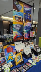 Vendor booth at Vintage Computer Festival Midwest displaying colorful art prints of vintage computers and gaming hardware.